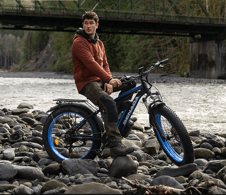 Man sitting on SENADA ARCHON PLUS fat tire electric bike by a rocky riverside, ideal for off-road adventures and long-distance riding.